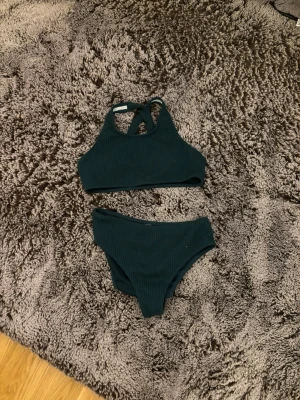 Svart ribbad bikini med hög midja - Stilren svart bikini med ribbat tyg. Toppen har hög hals och korsade axelband i ryggen. Underdelen är högt skuren och ger en modern look. Perfekt för stranden eller poolen.