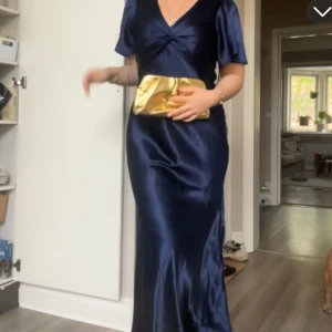 Marinblå långklänning i satin - Säljer en elegant marinblå långklänning i glansigt satinmaterial. Klänningen har korta ärmar, v-ringning och en snygg twistdetalj framtill. Perfekt för speciella tillfällen när du vill känna dig extra fin.