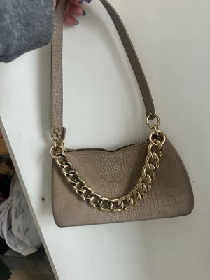 Beige handväska med guldkedja - Stilren beige handväska i krokopräglat material med en chunky guldfärgad kedjedetalj framtill. Väskan har ett kort axelband och stängs med dragkedja. Perfekt accessoar för att lyfta din outfit.