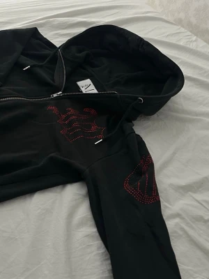 Svart hoodie med röd strasstext - Svart ziphoodie från MNML med huva och dragsko. På bröstet och ena ärmen finns stora röda bokstäver i strass som ger en snygg kontrast. Hoodien har fickor framtill och muddar vid ärmslut och nederkant. Perfekt för en streetwear-look.