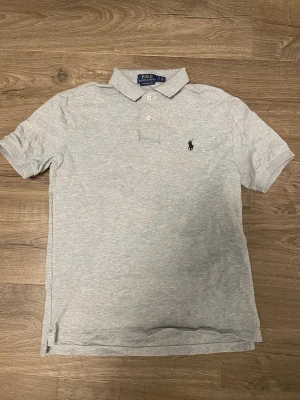Grå pikétröja från Polo Ralph Lauren - Klassisk ljusgrå pikétröja från Polo Ralph Lauren med korta ärmar och krage. 