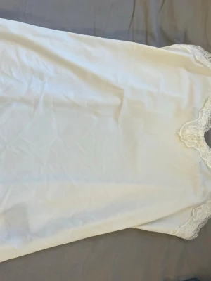 Vitt nattlinne med spetsdetaljer - Ett klassiskt vitt nattlinne med fina spetsdetaljer vid halsringningen och ärmsluten. Nattlinnet har en rak passform och är tillverkat i ett mjukt, tunt tyg som känns skönt mot huden. Perfekt för mysiga kvällar hemma.