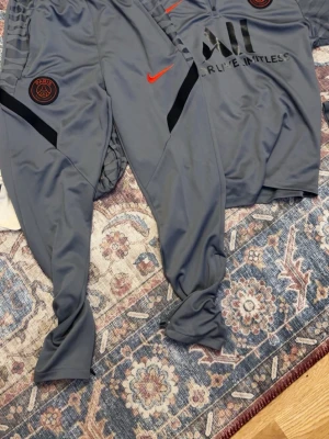 PSG träningsset i grått från Nike - Säljer ett grått träningsset från Nike och PSG med långärmad tröja och byxor. Setet har svarta och röda detaljer, PSG-logga och Nike Swoosh. Materialet är mjukt och stretchigt, perfekt för fotbollsträning eller gym.