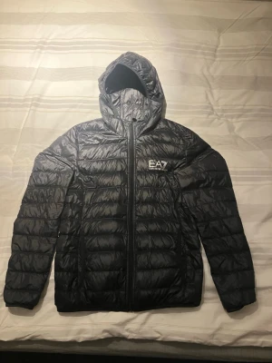 Svart dunjacka från EA7 Emporio Armani - Snygg svart dunjacka från EA7 Emporio Armani med huva och dragkedja framtill. Jackan har quiltad design och logga på bröstet. Perfekt för kyliga och varmare dagar och enkel att matcha med olika outfits. Fast pris, använd två gånger.