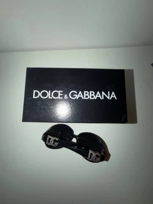 Dolce & Gabbana svarta solglasögon - Stilrena svarta solglasögon från Dolce & Gabbana med stora DG-loggor i silver och strass på skalmarna. Glasögonen har en klassisk rundad form och kommer med originalfodral och ask. Perfekt accessoar för en trendig look.