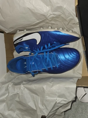 Blå Nike Tiempo fotbollsskor - Snygga blå Nike Tiempo fotbollsskor med vit sula och vit Nike-logga på sidan. Skorna har snörning och är designade för att ge bra grepp på planen. Perfekta för dig som vill sticka ut med stil på fotbollsplanen.