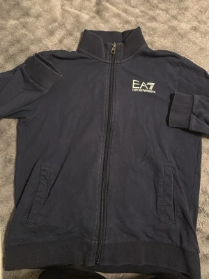 Marinblå ziptröja från EA7 - Snygg marinblå ziptröja från EA7 Emporio Armani med hög krage och vit logga på bröstet. Tröjan har dragkedja framtill, två fickor och ribbade muddar. Perfekt för en sportig och stilren look.