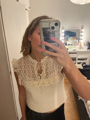 Beige stickad blus med fina detaljer - Supersöt beige blus med volangdetaljer vid axlarna. Blusen har knyt framtill vid halsen och är ärmlös. Storlek M men skulle snarare säga att den passar S eller XS.