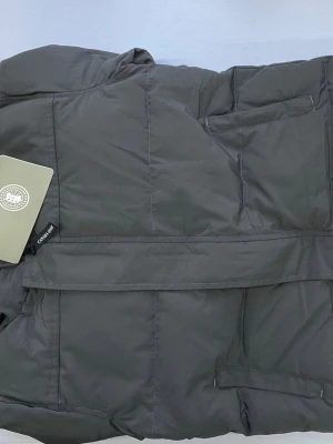 Grå pufferjacka från Canada Goose - Snygg grå pufferjacka från Canada Goose med quiltad design och hög krage. Jackan har diskret logotyp på ärmen och praktiska fickor framtill. Perfekt för kalla dagar och har en modern, stilren look.