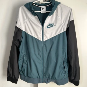 Vindjacka från Nike i blå, vit, svart - Snygg vindjacka från Nike i en kombination av blått, vitt och svart. Jackan har en dragkedja framtill och en huva för extra skydd. Perfekt för blåsiga dagar och har en sportig look med Nike-loggan på bröstet. 