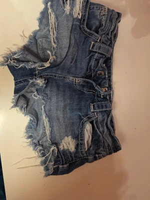 Blå slitna jeansshorts med fransar - Säljer ett par blå jeansshorts med råa kanter och slitningar för en edgy look. Shortsen har fem fickor, knappgylf och fransiga detaljer längs benen. Perfekta för sommaren och ger en avslappnad streetstyle-vibe.