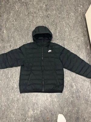 Svart pufferjacka från Nike med huva - Snygg svart pufferjacka från Nike med huva och dragkedja framtill. Jackan har quiltad design, två sidofickor och Nike-logga på bröstet. Perfekt för kalla dagar när du vill hålla stilen och värmen.