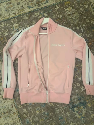 Rosa kofta från Palm Angels - Säljer en snygg rosa kofta från Palm Angels med vita och svarta ränder längs ärmarna. Koftan har dragkedja framtill, två fickor med dragkedja och en hög krage. Äkthetsbevis finns! Denna färgen är väldigt unik och svår att få tag i.