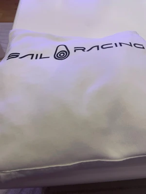 Vit hoodie från Sail Racing - Vit hoodie från Sail Racing med stor logga tryckt över bröstet. Klassisk modell med mjukt material och avslappnad passform. Perfekt för dig som gillar stilrena och sportiga plagg.