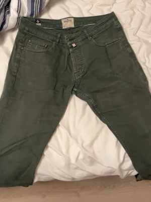 Gröna jeans från Morris Atelier - Snygga gröna jeans från Morris Atelier med klassisk femficksmodell och diskret logga på fickan. Jeansen har normal passform och är tillverkade i mjuk bomullsdenim. Perfekta för dig som vill ha en stilren och avslappnad look.