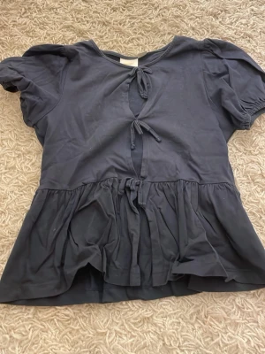 Svart blus med volang - Svart blus med korta puffärmar, tre knytningar framtill och volang nertill. Tillverkad i mjukt bomullstyg och har en avslappnad passform. Perfekt för en trendig och bekväm look.