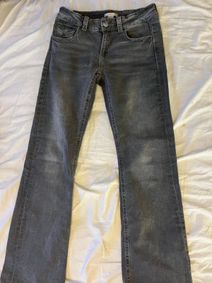 Grå bootcut jeans med detaljer - Säljer ett par gråa bootcut jeans med snygga detaljer på bakfickorna. Jeansen har normal passform och klassisk femficksdesign. Materialet är mjukt jeans-tyg som sitter skönt hela dagen. Perfekta för dig som gillar en avslappnad men ändå stilren look. Byxorna är k storlek 170. Hör av er vid frågor!☺️