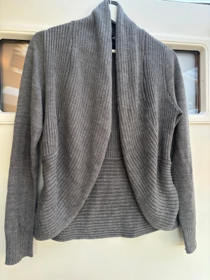 Grå ribbstickad kofta från H&M - Mysig grå kofta från H&M i ribbstickat material. Koftan har en öppen front utan knappar, lång ärm och en avslappnad passform. Perfekt att slänga över en t-shirt eller topp för extra värme. Enkel och stilren design som passar till det mesta.