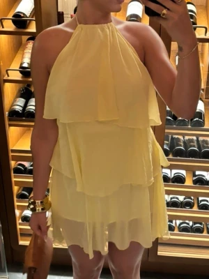 Gul volangklänning med halterneck + öppen rygg - Gul kortklänning med halterneck och flera lager volanger. Klänningen är ärmlös och har en luftig passform. Öppen rygg. Ett underlag under själva klänningen. Använd endast 2 gånger.