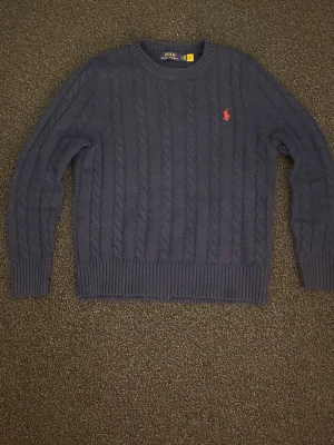 Mörkblå kabelstickad tröja Polo Ralph Lauren - Kabelstickad mörkblå tröja från Polo Ralph Lauren med klassisk röd broderad logga på bröstet. Tröjan har rund halsringning, ribbade muddar och nederkant. Perfekt för dig som gillar tidlös och stilren design.