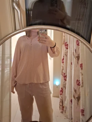 Ljus rosa blus - Mysig blus i ljusrosa med långärmad topp. Toppen har avslappnad passform, rundad halsringning med små knappar och lätt puffade ärmslut. Materialet känns mjukt och luftigt, perfekt för chill dagar fast endå uppklädd