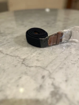 Mörkblått flätat bälte med skinn-detalj - Snyggt mörkblått flätat bälte med bruna skinn-detaljer och silverfärgat spänne. Perfekt för att lyfta vilken outfit som helst och funkar till både jeans och kostymbyxor. Klassisk design som passar till det mesta.