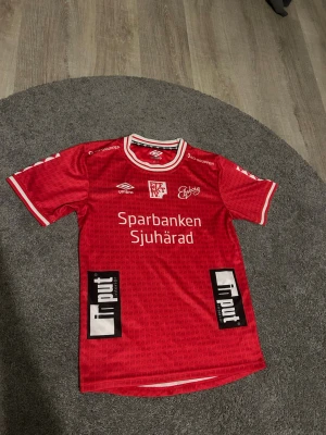 Röd matchtröja från Umbro - Säljer en röd matchtröja från Umbro med vita detaljer och tryck från Sparbanken Sjuhärad och Input. Tröjan har klubbmärke, sponsorloggor och rund hals med vit och röd kant. Perfekt för fotboll eller träning.obs har nedvänt en gång