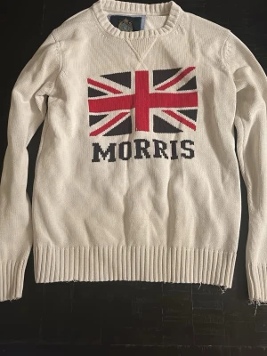 Vit stickad tröja Morris Union Jack - Vit stickad tröja från Morris med stor Union Jack-flagga i rött, blått och vitt på bröstet samt texten 'MORRIS' intill flaggan. Tröjan har rund halsringning och ribbade muddar vid ärmslut och nederkant. Perfekt för dig som gillar klassisk brittisk stil.
