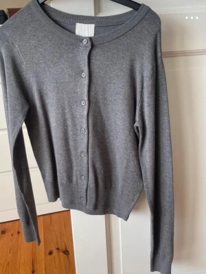 Grå basic kofta med rund hals - En enkel och stilren grå kofta med rund halsringning och knappar framtill. Långärmad modell i mjukt, tunt stickat material som passar perfekt till lager på lager. Ribbstickade muddar vid ärmslut och nederkant för en snygg passform.