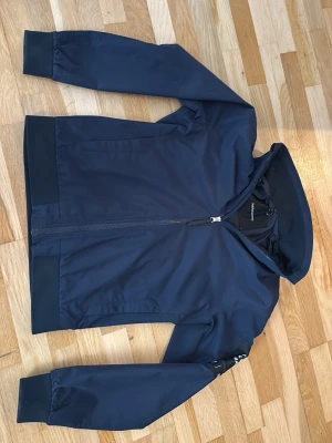 Marinblå vindjacka från Peak performance  - Marinblå vindjacka från Peak performance med  dragkedja framtill, ribbade muddar och diskret logga på ärmen. Jackan har en sportig och stilren look, perfekt för blåsiga dagar. Tillverkad i polyester och har normal passform. Och är i ett väldigt bra skick! Ny pris 2200 kr.