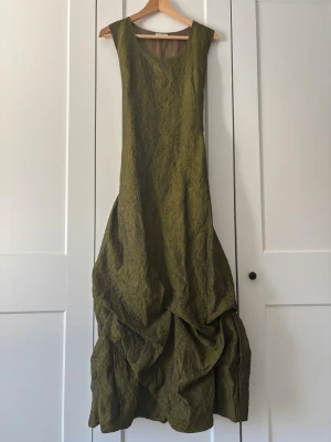 Olivgrön långklänning med snörning - Olivgrön långklänning med skrynklat tyg och draperad nederdel. Klänningen är ärmlös och har snörning i ryggen vilket gör att den passar flera storlekar.