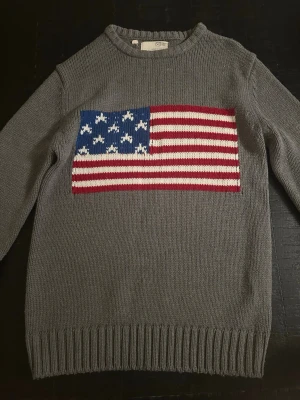 Grå stickad tröja med USA-flagga - Snygg grå stickad tröja från Ralph Lauren med en stor amerikansk flagga i rött, vitt och blått på bröstet. Tröjan har rund halsringning och ribbstickade muddar vid ärmslut och nederkant. Perfekt för dig som gillar klassisk preppy stil.