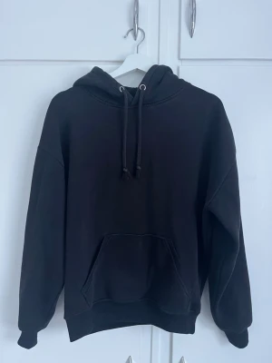 Svart hoodie med magficka - En klassisk svart hoodie med huva och dragsnören. Hoodien har en stor magficka framtill och ribbade muddar vid ärmslut och nederkant. Perfekt för en avslappnad och stilren look. Materialet känns mjukt och bekvämt.