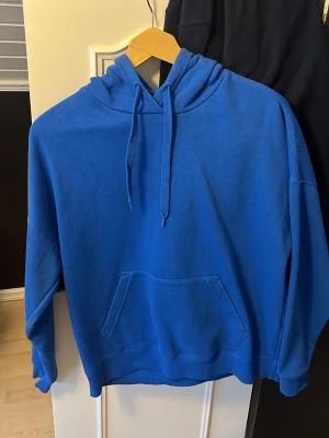 Blå hoodie med huva och magficka - En klassisk blå hoodie med huva och dragsko samt en stor magficka framtill. Hoodien har ribbade muddar vid ärmslut och nederkant. Perfekt för en avslappnad och stilren look. Materialet känns mjukt och bekvämt, troligen bomullsblandning.