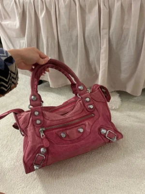Rosa Balenciaga handväska i skinn - Cool rosa handväska från Balenciaga i mjukt skinn med silverfärgade nitar och detaljer. Väskan har dubbla handtag, dragkedja framtill och en klassisk, lite slouchy form. Perfekt för dig som vill ha en statementväska med edgy vibe.