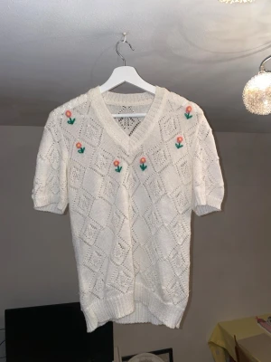 Vit stickad kortärmad tröja med blommor - Vit stickad tröja med korta ärmar och v-ringad hals. Tröjan har ett hålmönster i rutigt och är dekorerad med broderade blommor i rosa och gul med gröna blad. Jag hittar ingen lapp, så jag misstänker att den är hemmagjord. Finns därför ingen storlek, men cirka storlek M!