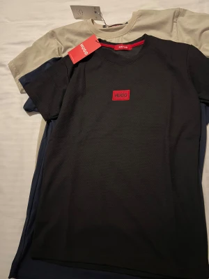 Svart HUGO t-shirt med röd logga - Snygg svart t-shirt från HUGO med röd logotyp på bröstet och röd HUGO-text på ärmen. Klassisk rund hals och korta ärmar. Materialet känns mjukt och har en lätt struktur. Perfekt för dig som gillar stilrena och moderna plagg.