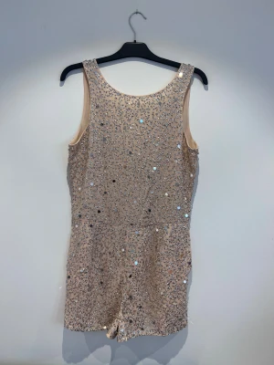 Beige paljettprydd byxdress - Snygg beige byxdress med massor av glittrande paljetter och stenar över hela plagget. Modellen är ärmlös med rund hals framtill och djup v-ringning bak. Materialet känns mjukt och har en lyxig känsla, perfekt för att sticka ut. Dragkedja i ryggen för enkel påklädning.