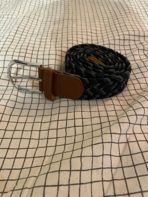 Mörkblå flätad bälte med bruna detaljer - Snyggt flätat bälte i mörkblå och grå toner med bruna skinnliknande detaljer vid spännet och änden. Spännet är i silverfärgad metall och bältet har en cool, vävd look som passar till jeans eller chinos.