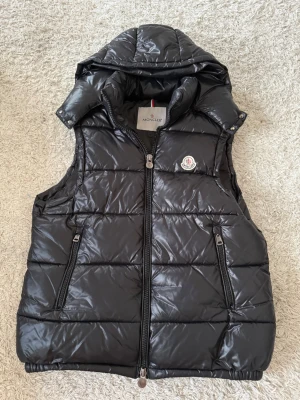 Svart dunväst från Moncler - Säljer en svart, quiltad dunväst från Moncler med glansig finish och avtagbar huva. Västen har två fickor med dragkedja och klassisk Moncler-logga på bröstet. Perfekt för kyliga dagar och har en cool, sportig vibe.