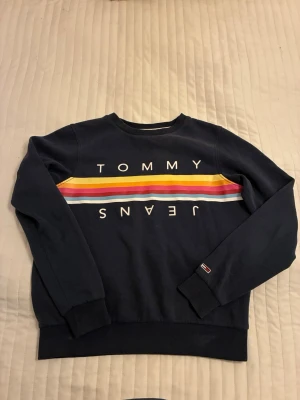 Mörkblå sweatshirt Tommy Jeans XS - Snygg mörkblå sweatshirt från Tommy hilfiger jeans i storlek XS. Tröjan har färgglada ränder i gult, rött, rosa och vitt. Rund hals och långa ärmar med ribbade muddar. Perfekt för dig som gillar streetwear och färg.Katt finns i huset.