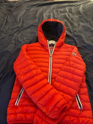 Röd pufferjacka från Colmar - OBS: Den har ett blått märke på ärmen men man märker inte av den.                 Säljer en röd pufferjacka från Colmar med huva och två sidofickor med dragkedja. Jackan har ett quiltat mönster och en snygg, sportig look. Perfekt för dig som gillar färg och vill sticka ut lite extra. Passar dig som vill ha en varm jacka med cool design.