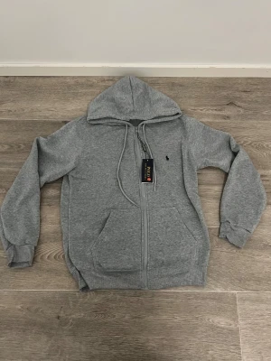 Grå mjukisbyxor från Polo Ralph Lauren - Snygga grå mjukisbyxor från Polo Ralph Lauren med dragsko i midjan och ribbade muddar vid bensluten. Byxorna har en klassisk logga broderad på ena benet och är gjorda i mjuk bomullsblandning för maxad komfort.