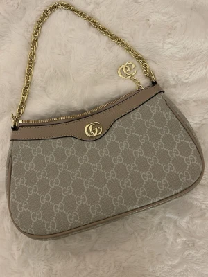 Beige axelväska med kedja - Säljer en beige axelväska från med monogram över hela väskan. Väskan har en snygg guldfärgad kedja som axelrem och guldfärgade detaljer, samt dragkedja upptill. Perfekt för dig som gillar lyxiga accessoarer.