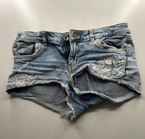 Blå jeansshorts med broderi  - Säljer ett par ljusblå jeansshorts med råa kanter och snyggt vitt blommigt broderi på framsidan. Shortsen har klassisk femficksdesign, knapp och dragkedja framtill samt slitna detaljer för en cool look. Passar även xxs och s