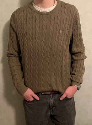 Olivgrön stickad tröja från Morris - Snygg olivgrön stickad tröja från Morris med klassiskt kabelstickat mönster och rund hals. Tröjan har långa ärmar och en liten broderad logga på bröstet. Storlek är XL men passar även perfekt till dig som bär L