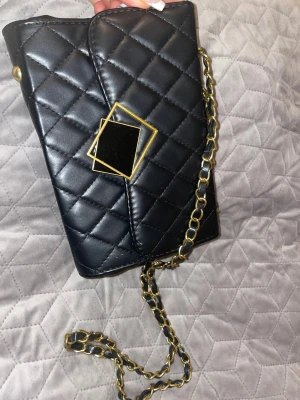 Svart quiltad axelväska med kedja - Snygg svart axelväska med quiltat mönster och guldfärgad kedja som axelrem. Väskan har en unik geometrisk detalj på locket och insidan är röd med dragkedjefack. Perfekt för dig som gillar stilrena accessoarer med lite edge.