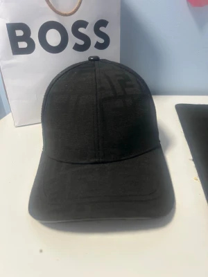 Svart Fendi keps med FF-mönster(Utan påse) - Snygg svart keps från Fendi med diskret FF-logomönster över hela ytan. Klassisk böjd skärm och justerbart spänne bak i svart läder med gulddetalj. Kepsen är tillverkad i ett slitstarkt tyg och har Fendi-logga på insidan. Perfekt för dig som gillar exklusiva accessoarer.