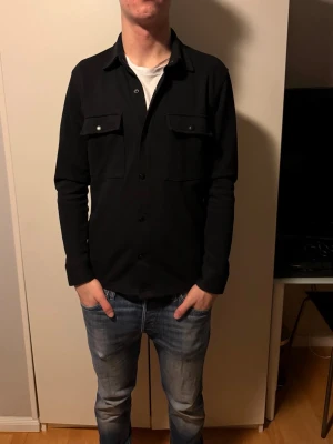 Svart overshirt från Selected Homme - Svart overshirt från Selected Homme i stilren design med klassisk krage och två stora bröstfickor med lock och knapp. Jackan har knäppning framtill och långärmade ärmar. Perfekt för dig som gillar minimalistisk och clean stil.