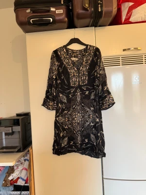 Svart kortklänning med mönster - Supersnygg svart kortklänning från Odd Molly med ljusa och beige detaljer i ett unikt blommigt och paisley-inspirerat mönster. Klänningen har v-ringning, trekvartsärmar med volang och är gjord i mjuk bomull. Perfekt för dig som gillar boho vibes och coola prints. Från märket Odd Molly. Storlek 2 som enligt deras storleksguide motsvarar en M. Mycket fint skick, jag hittar inga synliga anmärkningar eller defekter.  